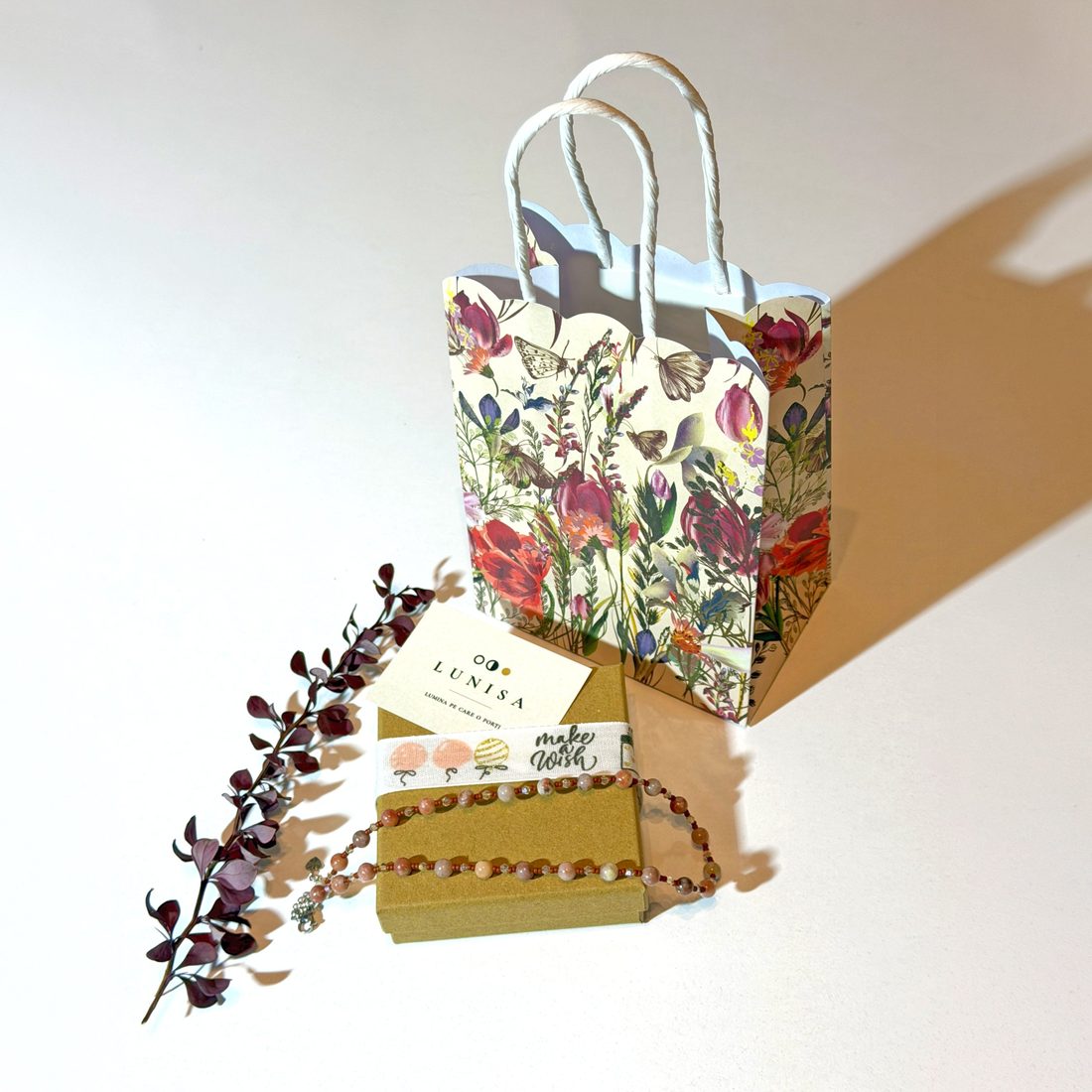 Sacoșă cadou florală, cutie kraft cu panglică și colier, lângă o crenguță